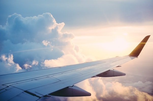 The view out of a plane window. The plane wing is in the foreground framed by clouds and the sun on the horizon.